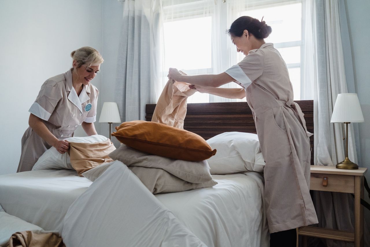 team-01 Two housekeepers in uniform fixing a hotel room bed with fresh linen and pillows.